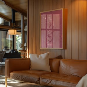 May include: A rectangular framed wall art piece featuring a deep pink background with a dark, slender botanical silhouette of a plant stem. The artwork is mounted on a vertical wood-paneled wall above a tan leather sofa with a white throw pillow.