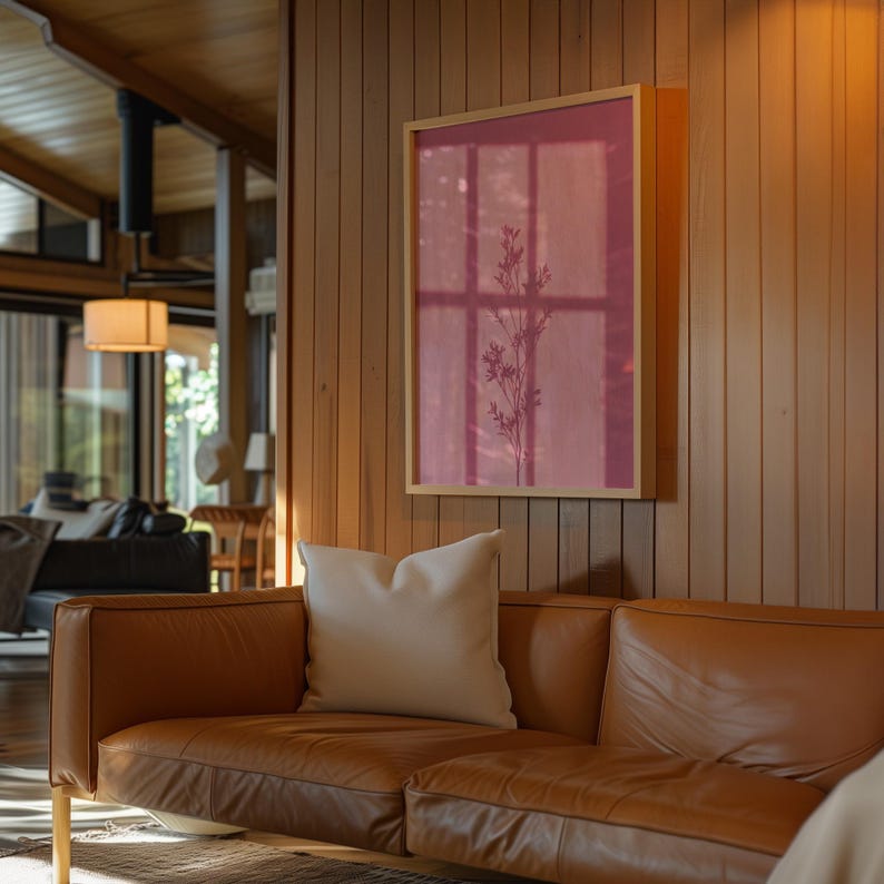 May include: A rectangular framed wall art piece featuring a deep pink background with a dark, slender botanical silhouette of a plant stem. The artwork is mounted on a vertical wood-paneled wall above a tan leather sofa with a white throw pillow.