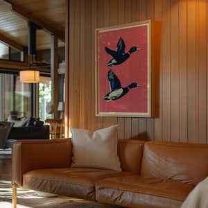 May include: A framed art print featuring two black ducks in flight against a red background. The ducks have white and blue accents. The artwork is displayed on a wooden wall above a brown leather sofa.