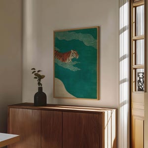May include: A framed art print featuring a tiger swimming in a teal-colored sea with white wave accents. The tiger has orange and black stripes. The artwork is in a wooden frame and is displayed on a wooden cabinet.