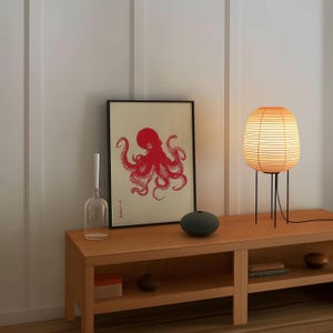 May include: A wooden console table with a framed red octopus print, a glass vase, and a small, round, dark green vase. A paper lantern lamp with a black metal base sits on the right. The lamp is lit, casting a warm glow.