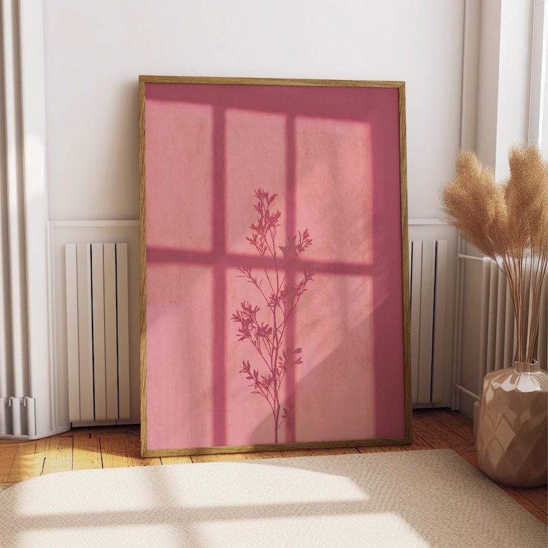 May include: A rectangular wall art print in a wooden frame featuring a solid dusty rose background with a dark silhouette of a slender plant stem with small leaves and flowers, overlaid by a grid-like shadow pattern from a window frame.