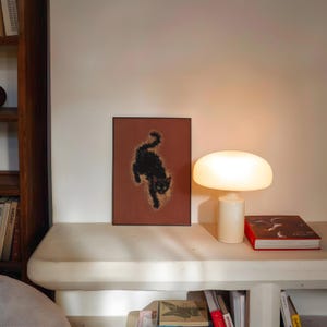 May include: A framed art print featuring a black cat silhouette on a reddish-brown background. A white mushroom-shaped lamp and a red book are on a white shelf. A wooden bookshelf is visible on the left.
