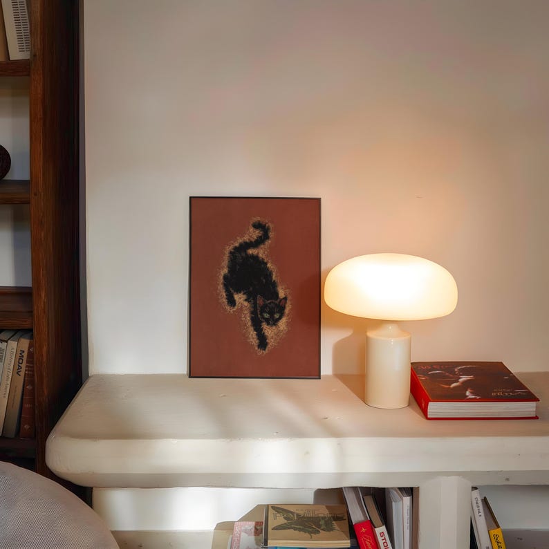 May include: A framed art print featuring a black cat silhouette on a reddish-brown background. A white mushroom-shaped lamp and a red book are on a white shelf. A wooden bookshelf is visible on the left.