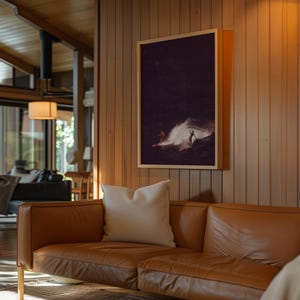 May include: A brown leather sofa with a white throw pillow in front of a wooden wall with a framed photograph of two people surfing in a dark ocean.