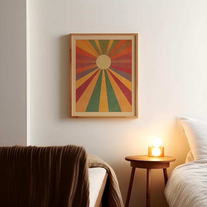 Op de afbeelding: Een ingelijst afdruk van een kleurrijk zonnestralenontwerp met een witte cirkel in het midden. De afdruk hangt aan een witte muur boven een houten nachtkastje met een lamp en een bed met een bruin dekbed.