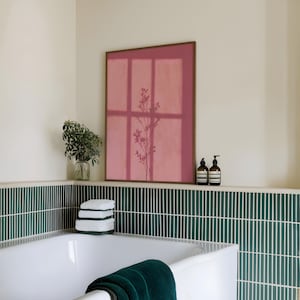 May include: A rectangular wall art print in a dark wood frame featuring a muted rose-pink background with a dark silhouette of a slender, branching plant. The artwork is displayed above a white bathtub with dark green vertical tile wall accents.