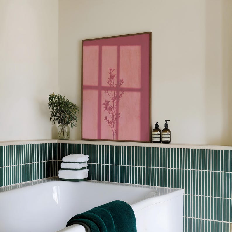 May include: A rectangular wall art print in a dark wood frame featuring a muted rose-pink background with a dark silhouette of a slender, branching plant. The artwork is displayed above a white bathtub with dark green vertical tile wall accents.