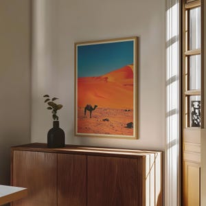 K&ouml;nnte beinhalten: Ein gerahmter Druck eines Kamels, das durch eine W&uuml;stenlandschaft geht. Die Sandd&uuml;nen sind in einem leuchtenden Orange gehalten, und der Himmel ist tiefblau. Das Kamel ist vor dem leuchtend orangenen Sand silhouettiert.