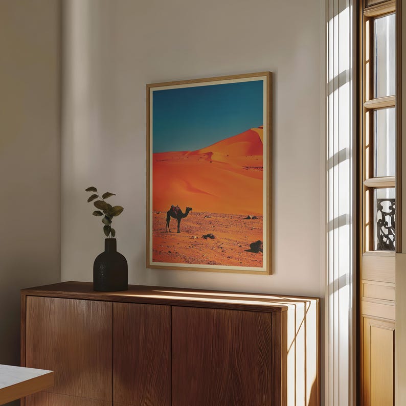 K&ouml;nnte beinhalten: Ein gerahmter Druck eines Kamels, das durch eine W&uuml;stenlandschaft geht. Die Sandd&uuml;nen sind in einem leuchtenden Orange gehalten, und der Himmel ist tiefblau. Das Kamel ist vor dem leuchtend orangenen Sand silhouettiert.