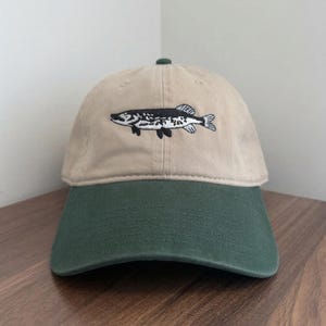 Könnte beinhalten: Beige und grüne Baseballkappe mit einem gestickten Fischdesign. Die Kappe hat eine beige Krone und einen grünen Schirm. Der Fisch ist schwarz und weiß und setzt einen Kontrast.