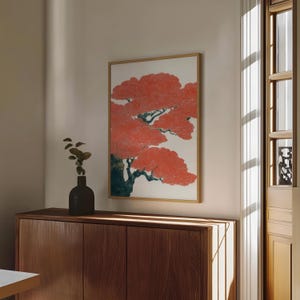 May include: A framed art print featuring a stylized tree with vibrant red foliage against a white background. The artwork is in a wooden frame and hangs on a neutral-colored wall. A dark vase with greenery sits on a wooden cabinet below.