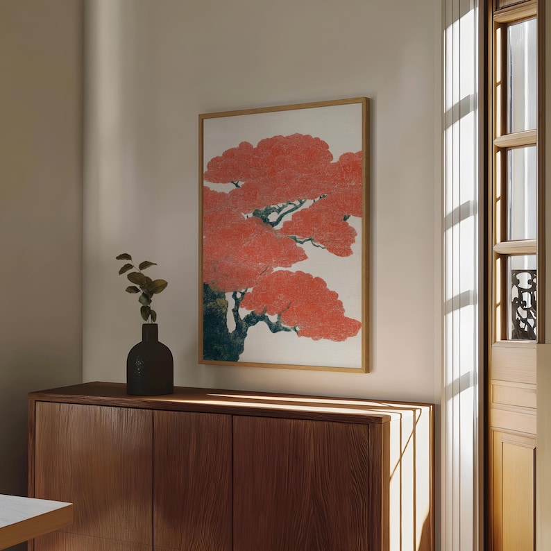May include: A framed art print featuring a stylized tree with vibrant red foliage against a white background. The artwork is in a wooden frame and hangs on a neutral-colored wall. A dark vase with greenery sits on a wooden cabinet below.