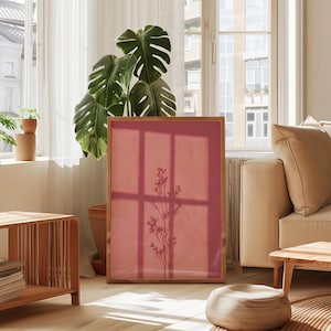 May include: A rectangular framed print featuring a solid dusty rose background with a dark silhouette of a slender plant stem with leaves centered in the middle. The frame is made of light-toned wood and stands upright on a light-colored floor.