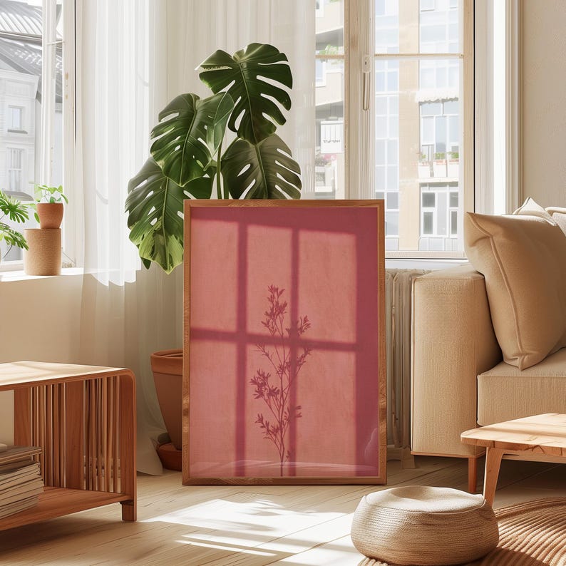 May include: A rectangular framed print featuring a solid dusty rose background with a dark silhouette of a slender plant stem with leaves centered in the middle. The frame is made of light-toned wood and stands upright on a light-colored floor.