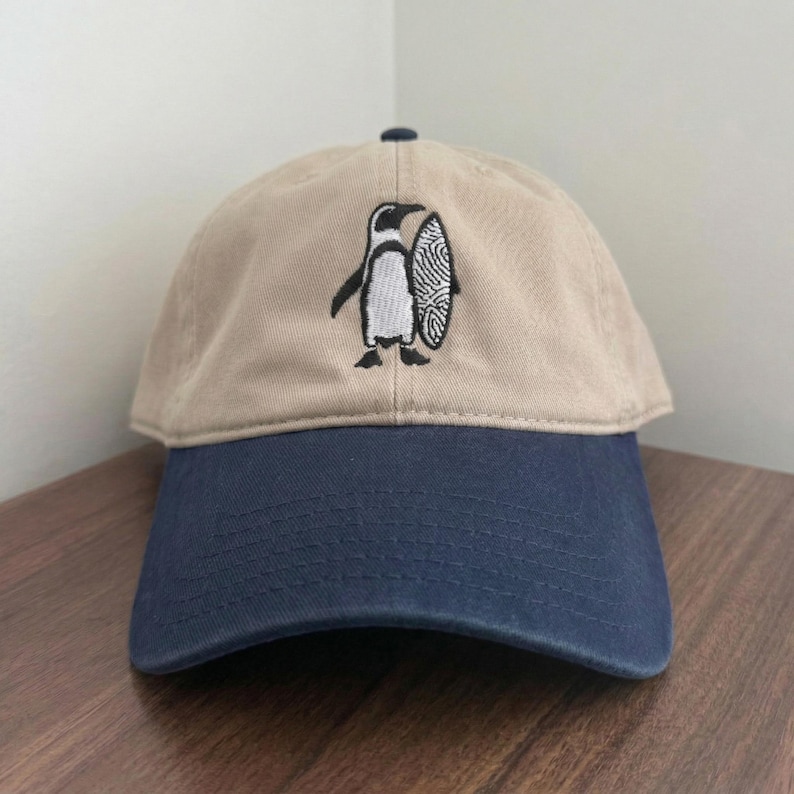 May include: A tan and navy blue baseball cap with a penguin and surfboard embroidered design. The cap features a curved brim and an adjustable strap. The penguin is black and white, holding a surfboard with a fingerprint design.