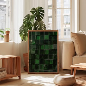 May include: A framed artwork featuring a mosaic of dark green glass squares. The artwork is set in a natural wood frame and leans against a wall. The room has a modern aesthetic with a large plant and neutral-toned furniture.
