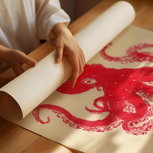 May include: A red octopus illustration on a cream-colored paper is being unrolled. The octopus design is detailed with visible tentacles and a central eye. The paper is on a wooden surface, and the unrolling reveals the artwork.