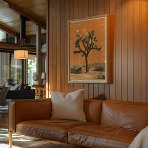 May include: A brown leather sofa with a white throw pillow in front of a wood-paneled wall. A framed print of a Joshua tree in a desert landscape hangs on the wall.