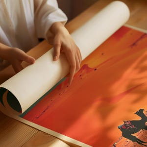 K&ouml;nnte beinhalten: Ein aufgerollter Druck eines W&uuml;stenlandschafts mit Kamelen im Vordergrund. Der Druck wird von einer Hand auf einer Holzoberfl&auml;che abgerollt.