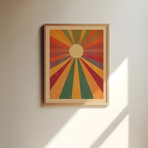 Op de afbeelding: Een ingelijste print van een kleurrijk zonnestralenontwerp met een witte cirkel in het midden. De achtergrond is lichtgeel met tinten rood, oranje, geel, groen en paars die vanuit het midden uitstralen.