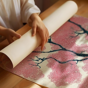 May include: A person is unrolling a scroll featuring a pink floral design with dark blue branches. The scroll is cream-colored and the floral pattern is on a white background. The scene is set on a wooden surface.