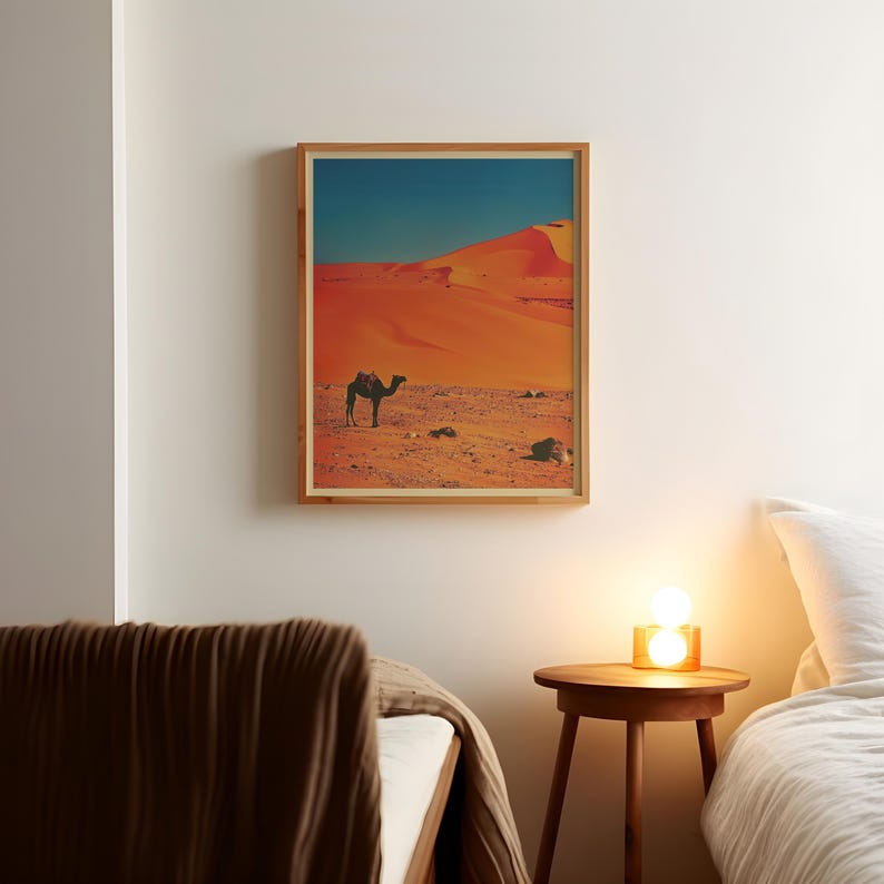 K&ouml;nnte beinhalten: Ein gerahmter Druck eines Kamels, das durch eine W&uuml;stenlandschaft geht. Die Sandd&uuml;nen sind in einem leuchtenden Orange gehalten, und der Himmel ist tiefblau. Das Kamel ist vor dem leuchtend orangenen Sand silhouettiert.