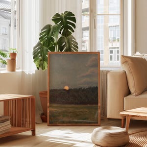 Puede incluir: Una pintura de paisaje enmarcada representa una puesta de sol sobre un bosque y un campo. La obra de arte est&aacute; en un marco de madera y se exhibe en una habitaci&oacute;n luminosa con una gran planta verde y un sof&aacute; beige. Los colores de la pintura incluyen tonos de naranja, verde y marr&oacute;n.