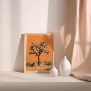 May include: A framed print of a Joshua tree in a desert landscape with an orange sky. The print is displayed on a white shelf with two white ceramic vases and dried pampas grass.