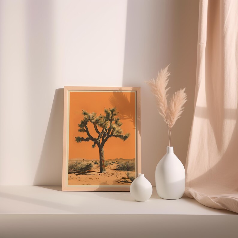 May include: A framed print of a Joshua tree in a desert landscape with an orange sky. The print is displayed on a white shelf with two white ceramic vases and dried pampas grass.