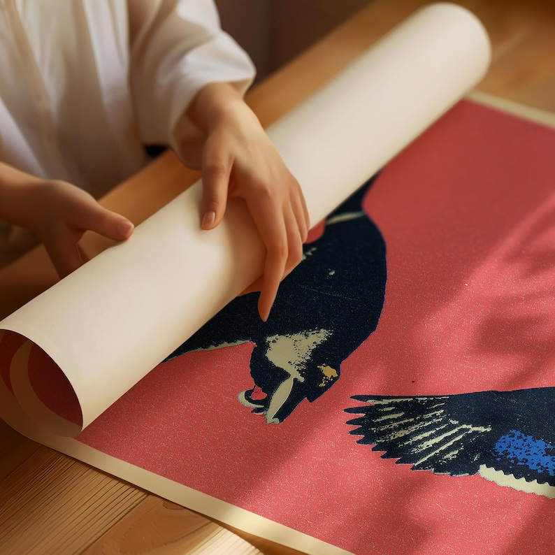 May include: A rolled-up art print featuring a bird design in shades of navy blue and white against a pink background. The print is being unrolled on a wooden surface, revealing the artwork. The print is rolled up and held by a person.