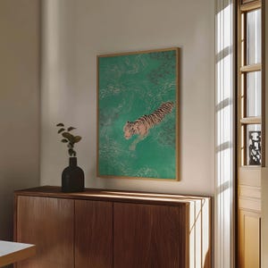 以下が含まれることがあります： 緑色の水の中を泳ぐトラを描いた額入りのアートプリント。トラはピンクと黒の縞模様です。アートワークは木製のフレームに入っており、白い壁に掛けられています。木製のキャビネットと黒い花瓶が前景にあります。