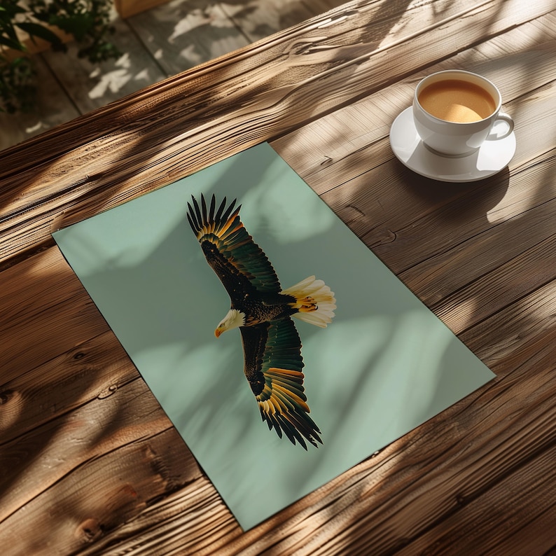 Pu&ograve; includere: Una stampa di un'aquila calva in volo, con le ali spiegate, su uno sfondo azzurro. Le piume dell'aquila sono dettagliate con sfumature di marrone, verde, giallo e bianco. Una tazza di caff&egrave; &egrave; su un piattino bianco su un tavolo di legno.