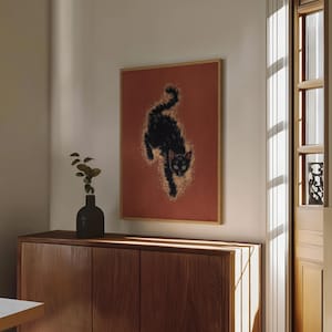 May include: A framed art print featuring a black cat with green eyes against a reddish-orange background. The cat is depicted in a dynamic pose, with a golden glow surrounding its form. The artwork is displayed in a room with natural light.
