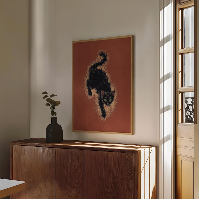 May include: A framed art print featuring a black cat with green eyes against a reddish-orange background. The cat is depicted in a dynamic pose, with a golden glow surrounding its form. The artwork is displayed in a room with natural light.