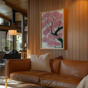 May include: A framed artwork depicting a pink flowering tree with a green trunk and branches. The artwork is mounted on a wooden wall above a brown leather sofa with a white pillow. The room has a warm, inviting atmosphere.