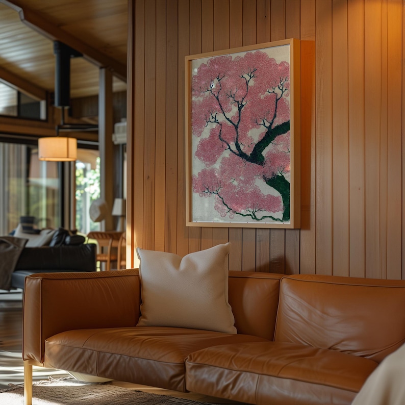 May include: A framed artwork depicting a pink flowering tree with a green trunk and branches. The artwork is mounted on a wooden wall above a brown leather sofa with a white pillow. The room has a warm, inviting atmosphere.