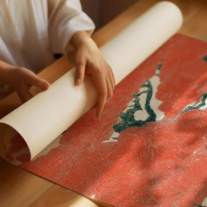 May include: A rolled-up, off-white paper print being unrolled onto a red and white patterned surface. The print features a design with red and white floral patterns and green accents. The scene is set on a wooden table.