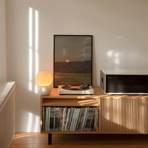 Puede incluir: Una consola de discos de madera clara con un tocadiscos blanco reproduciendo un disco de vinilo. Una pintura de paisaje enmarcada y una l&aacute;mpara esf&eacute;rica est&aacute;n encima. La consola tiene un armario con listones de madera verticales y una balda inferior llena de discos.