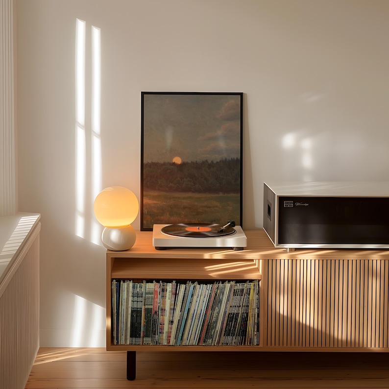 Puede incluir: Una consola de discos de madera clara con un tocadiscos blanco reproduciendo un disco de vinilo. Una pintura de paisaje enmarcada y una l&aacute;mpara esf&eacute;rica est&aacute;n encima. La consola tiene un armario con listones de madera verticales y una balda inferior llena de discos.