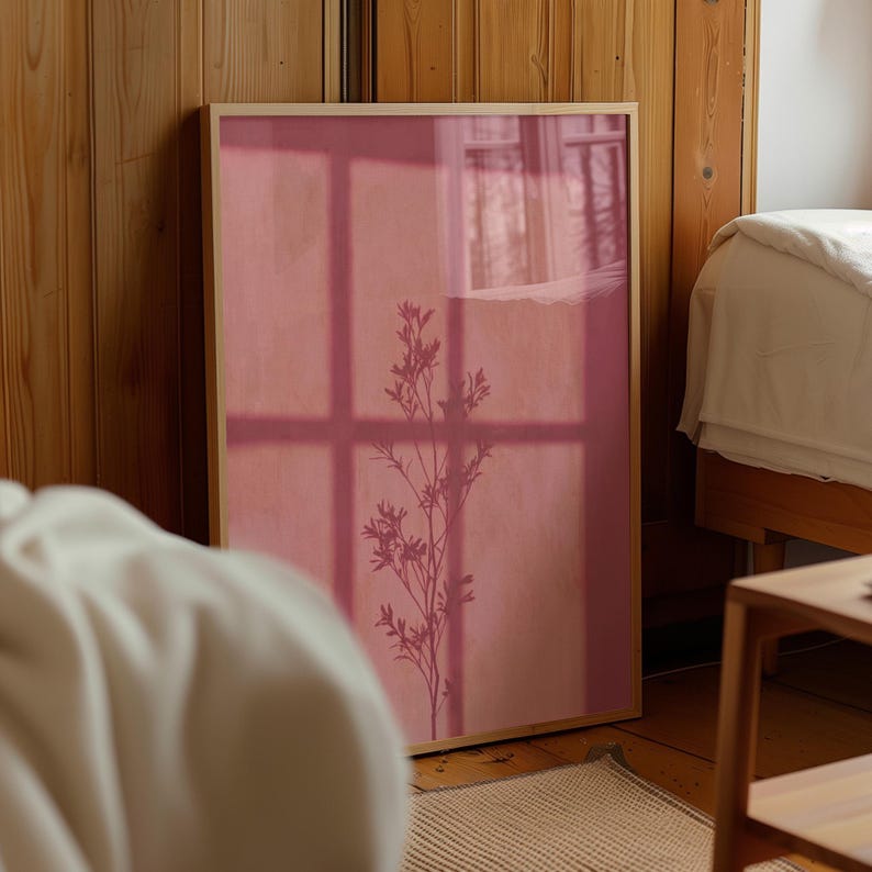 May include: A rectangular wall art print in a light wood frame featuring a solid dusty rose background with a dark silhouette of a slender plant stem with leaves and a grid-like shadow pattern cast across the surface of the artwork.
