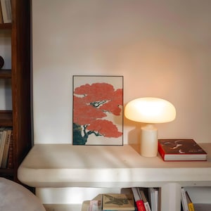 May include: A modern interior scene featuring a white mushroom-shaped table lamp, a framed artwork with a red tree design, and a red book. The lamp is on a white surface, with a wooden bookshelf visible on the left.