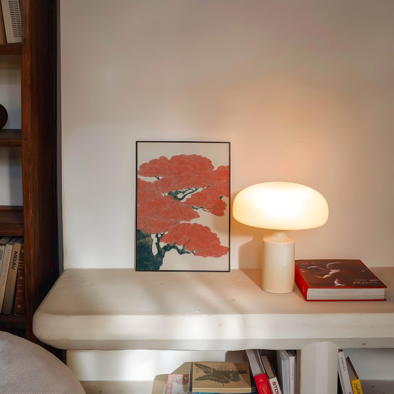 May include: A modern interior scene featuring a white mushroom-shaped table lamp, a framed artwork with a red tree design, and a red book. The lamp is on a white surface, with a wooden bookshelf visible on the left.