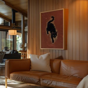 May include: A framed artwork featuring a black cat on a reddish-brown background hangs on a wooden wall. The cat is depicted in a dynamic pose, with a light-colored, textured effect around it. A brown leather sofa with a white pillow is in the foreground.