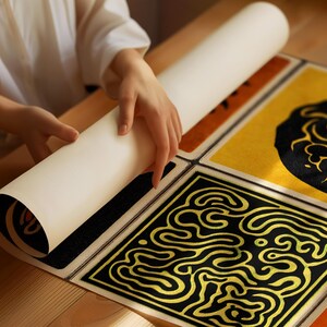 May include: A person unrolling a cream-colored paper scroll onto a set of four square mats. The mats feature black and gold geometric designs on a yellow and black background. The mats are on a wooden surface.