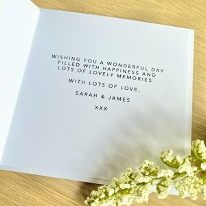 May include: A white greeting card with black printed text that reads: WISHING YOU A WONDERFUL DAY FILLED WITH HAPPINESS AND LOTS OF LOVELY MEMORIES. WITH LOTS OF LOVE, SARAH & JAMES XXX. The card is partially obscured by a sprig of small white flowers.