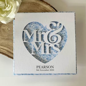 May include: A square white card featuring a heart-shaped cutout with the text Mr & Mrs. The background inside the heart is a blue and white floral pattern. Below the heart, the text PEARSON and 9th November 2024 are printed in black ink.