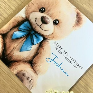 May include: A square greeting card featuring a watercolor illustration of a light brown teddy bear with a bright blue bow around its neck. The text reads: HAPPY 3RD BIRTHDAY TO OUR GORGEOUS SON Joshua in black and blue script lettering.