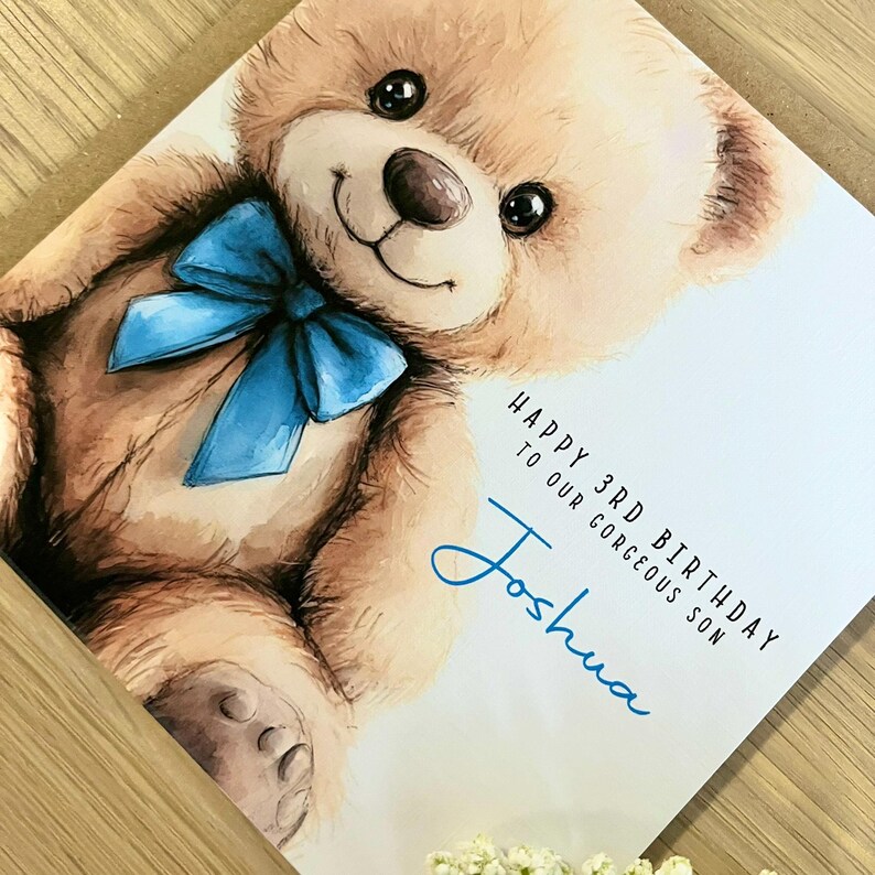 May include: A square greeting card featuring a watercolor illustration of a light brown teddy bear with a bright blue bow around its neck. The text reads: HAPPY 3RD BIRTHDAY TO OUR GORGEOUS SON Joshua in black and blue script lettering.