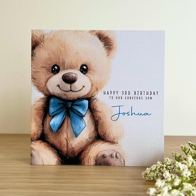 May include: A square greeting card featuring a watercolor illustration of a light brown teddy bear with a bright blue bow tie. The text reads: Happy 3rd Birthday to our gorgeous son, Joshua. The card has a textured white background.